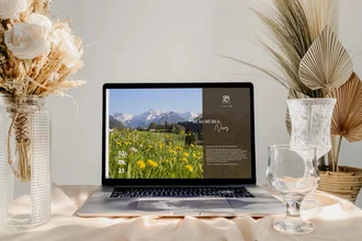 Modernes Laptop-Mockup mit Website der Burgmühle Gourmet & Spa, präsentiert in stilvoller Umgebung mit Trockenblumen und Gläsern – TYPO3 Webdesign für Hotel in den Alpen