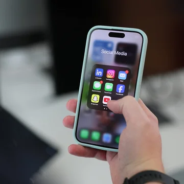 Hand hält ein Smartphone mit geöffnetem Social-Media-Ordner; sichtbar sind App-Icons wie LinkedIn, Instagram, Facebook, TikTok und YouTube.