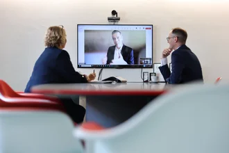 Videokonferenz bei Denkströme: Zwei Personen sitzen im Besprechungsraum am Tisch und sprechen über einen großen Bildschirm mit zugeschaltetem Gesprächspartner