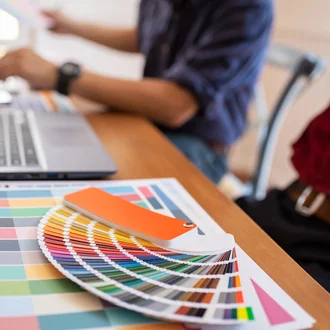 Farbfächer und Farbpaletten auf einem Schreibtisch mit Laptop im Hintergrund – kreative Arbeit im Bereich Grafikdesign und Branding